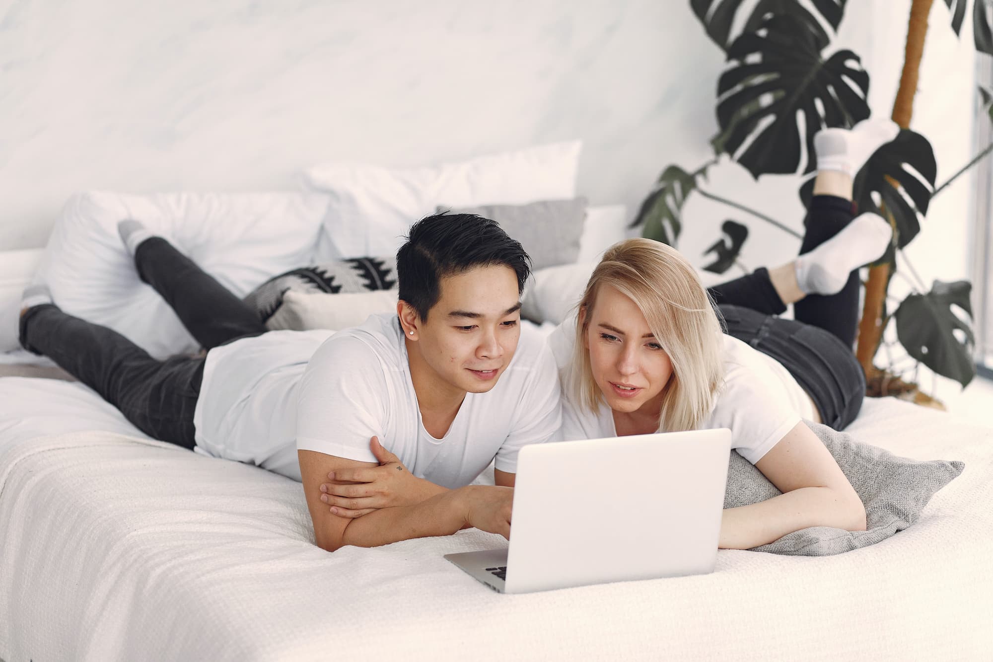 Interracial couple are lying on their bed looking at their laptop